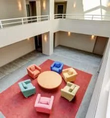 An apartment building community lounge with armchairs arranged around a round table on a red rug, seen from the balcony above