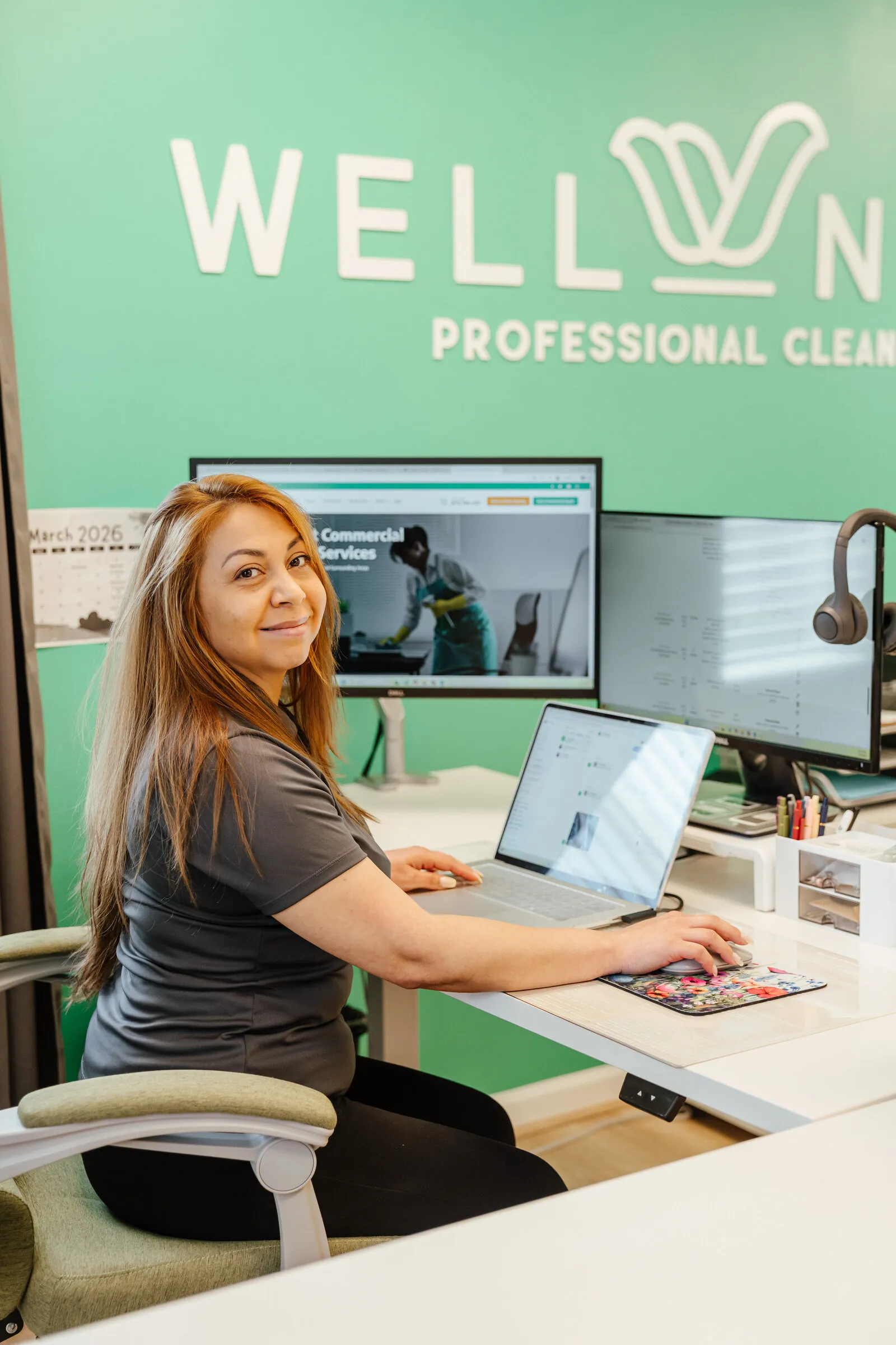 A WellNest team member wearing a headset at a desk, taking a call in front of the green WellNest office wall.