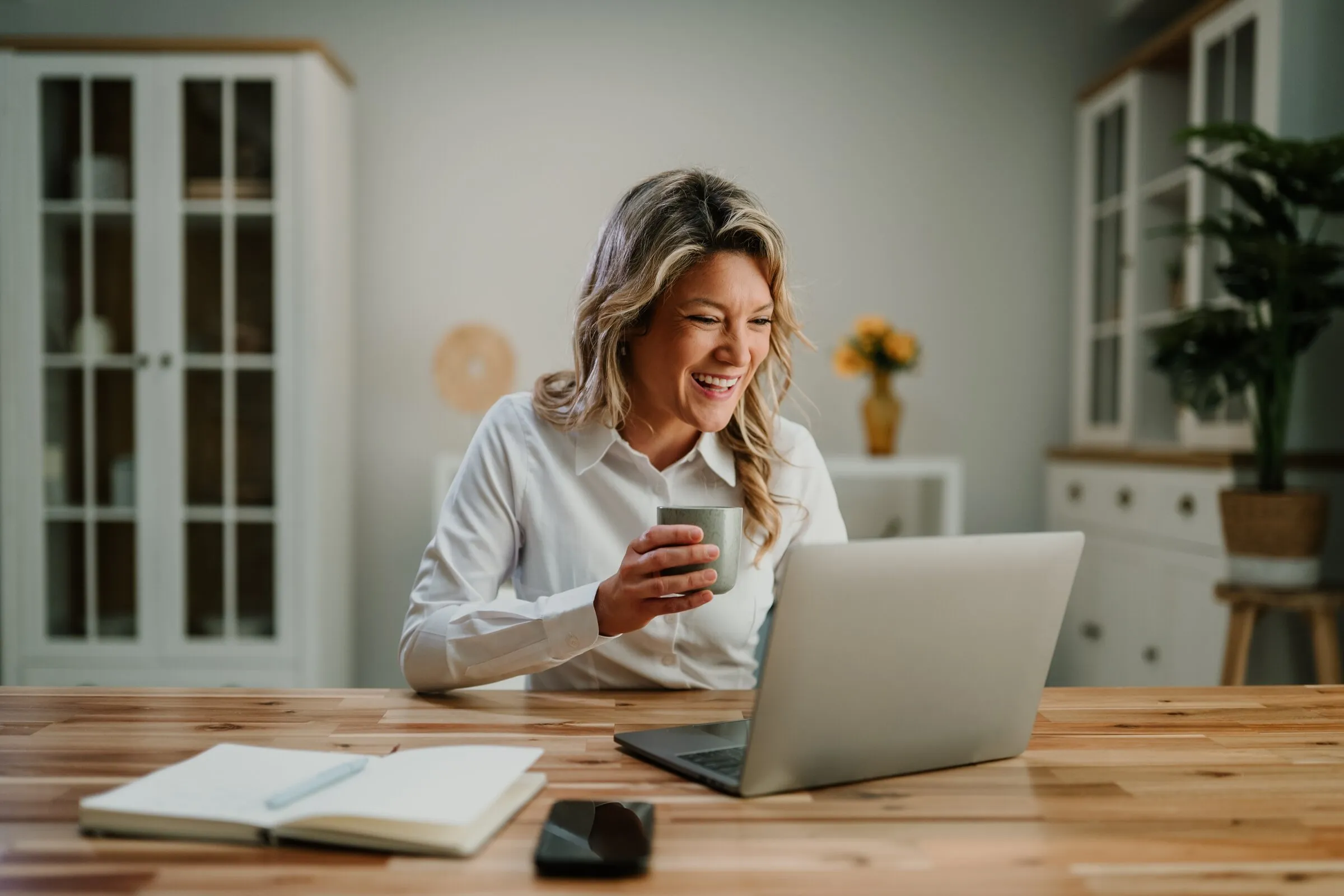A woman smiling at her phone beside an open laptop and notebook on a sunlit kitchen counter.