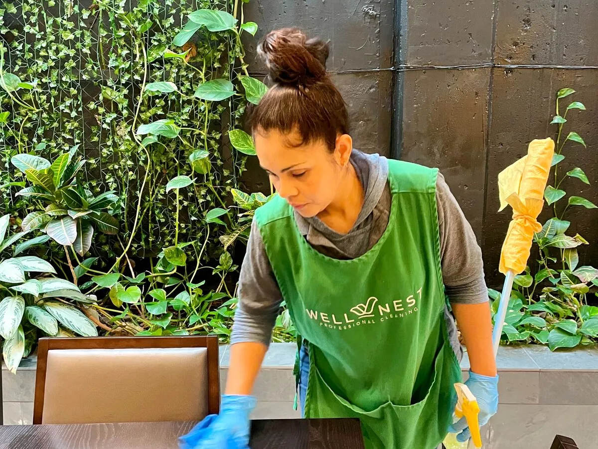WellNest cleaner in a green apron wiping down a conference room table during a commercial office cleaning
