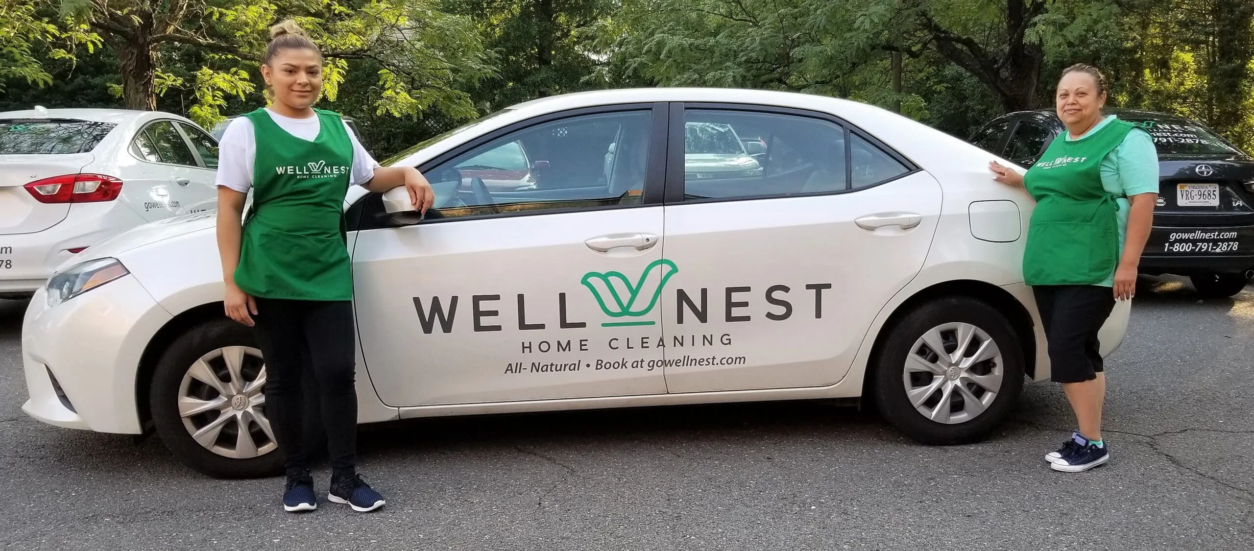 Two WellNest cleaners in green aprons standing on either side of a white WellNest company car in a shaded parking lot